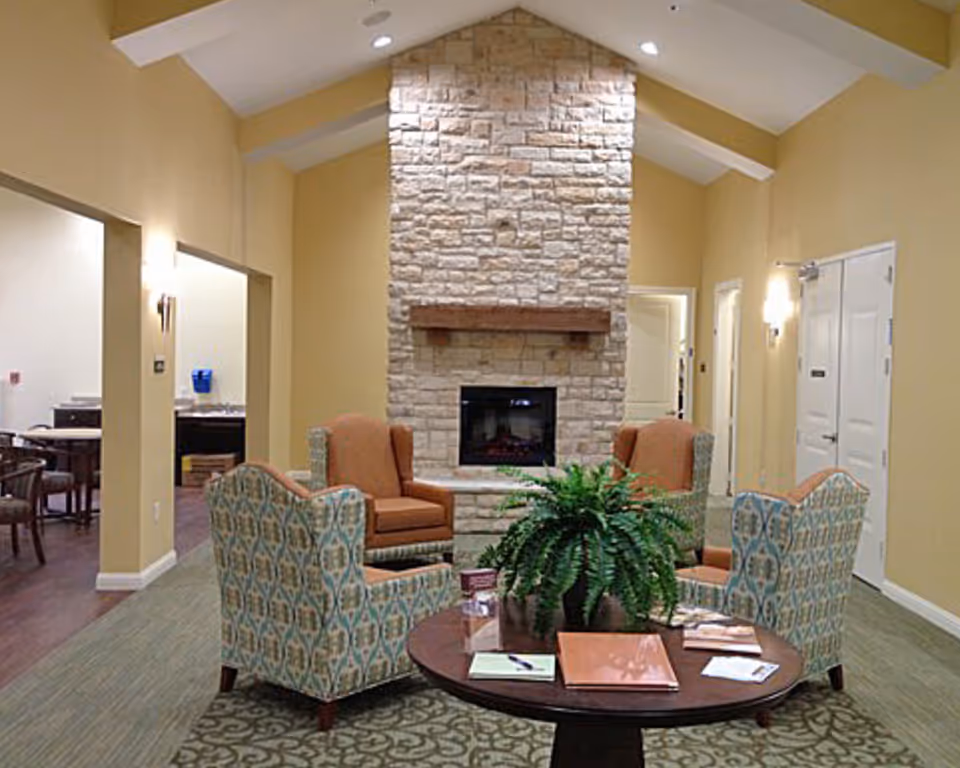 A cozy seating area with patterned armchairs around a round table and a stone fireplace in a senior living common room.