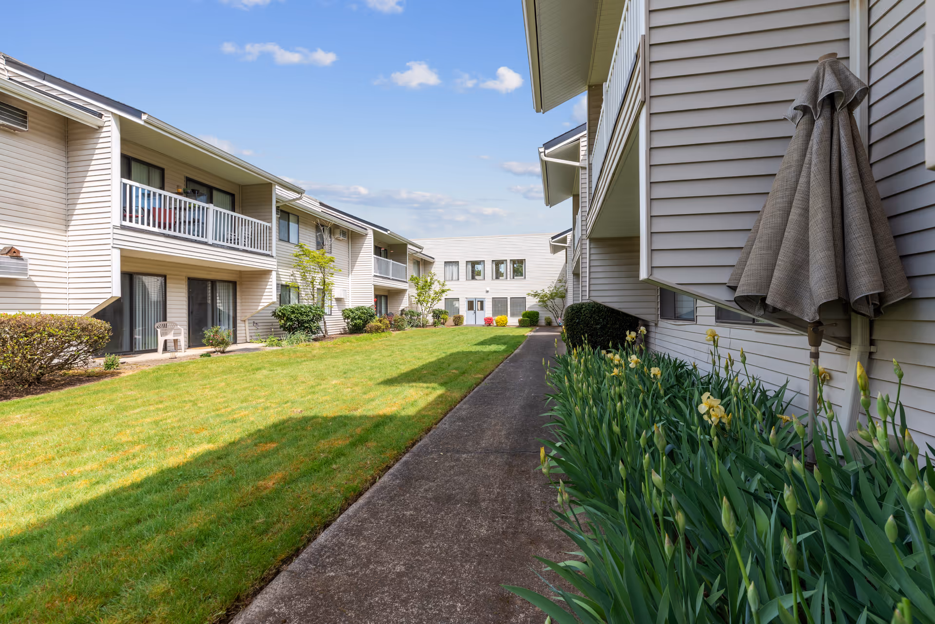 Outdoor courtyard area of Summerfield Estates Retirement Community featuring a green lawn, flower beds with blooming plants, a concrete walkway, and two-story beige buildings with balconies and sliding glass doors under a blue sky with scattered clouds.