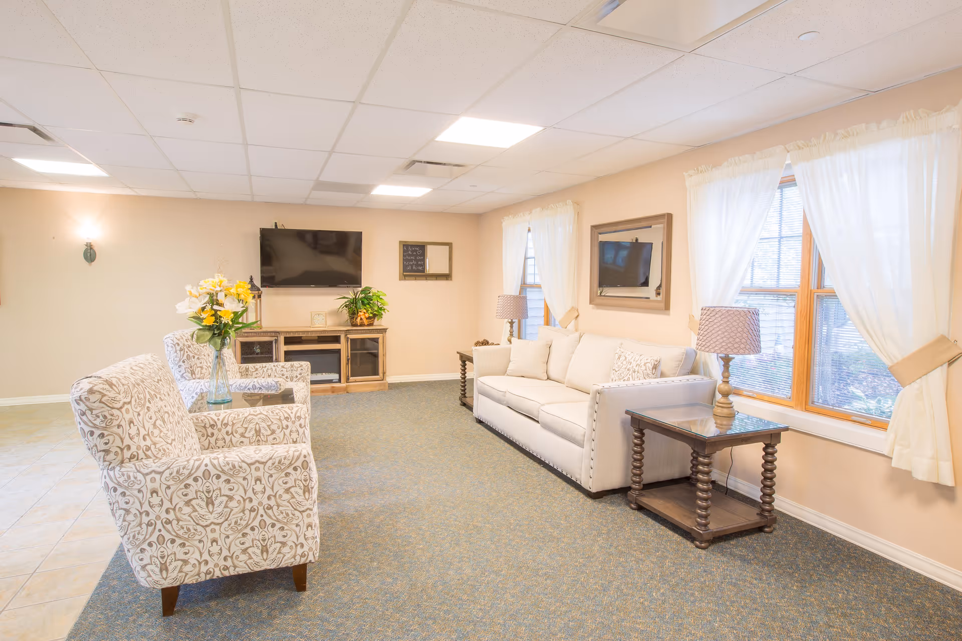 A bright and cozy living room with a beige sofa, two patterned armchairs, a wooden side table with a lamp, a TV mounted on the wall above a wooden cabinet, and windows with white curtains letting in natural light.