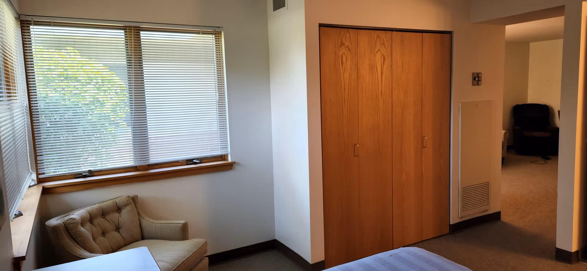 A corner of a room with a beige armchair under a window with closed blinds. Next to the chair is a wooden closet with four vertical doors. The room has light-colored walls and carpeted floor. In the background, there is a partial view of another room with a dark recliner chair.