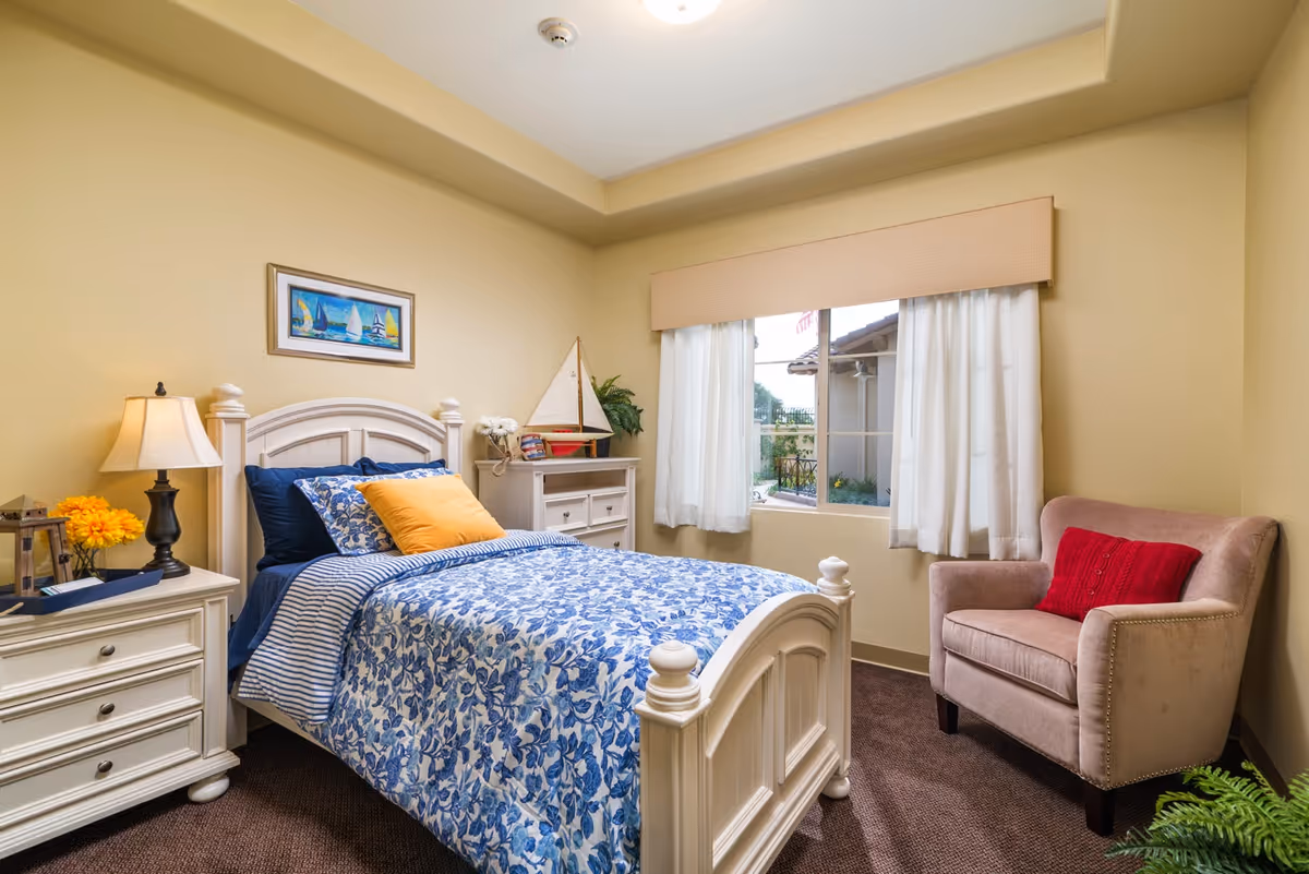 A cozy bedroom with a white wooden bed frame, blue and white floral bedding, and a yellow pillow. There is a nightstand with a lamp and flowers on the left side of the bed, a dresser with a decorative sailboat and a plant near the window, and a beige armchair with a red pillow on the right. The room has beige walls and a window with white curtains letting in natural light.