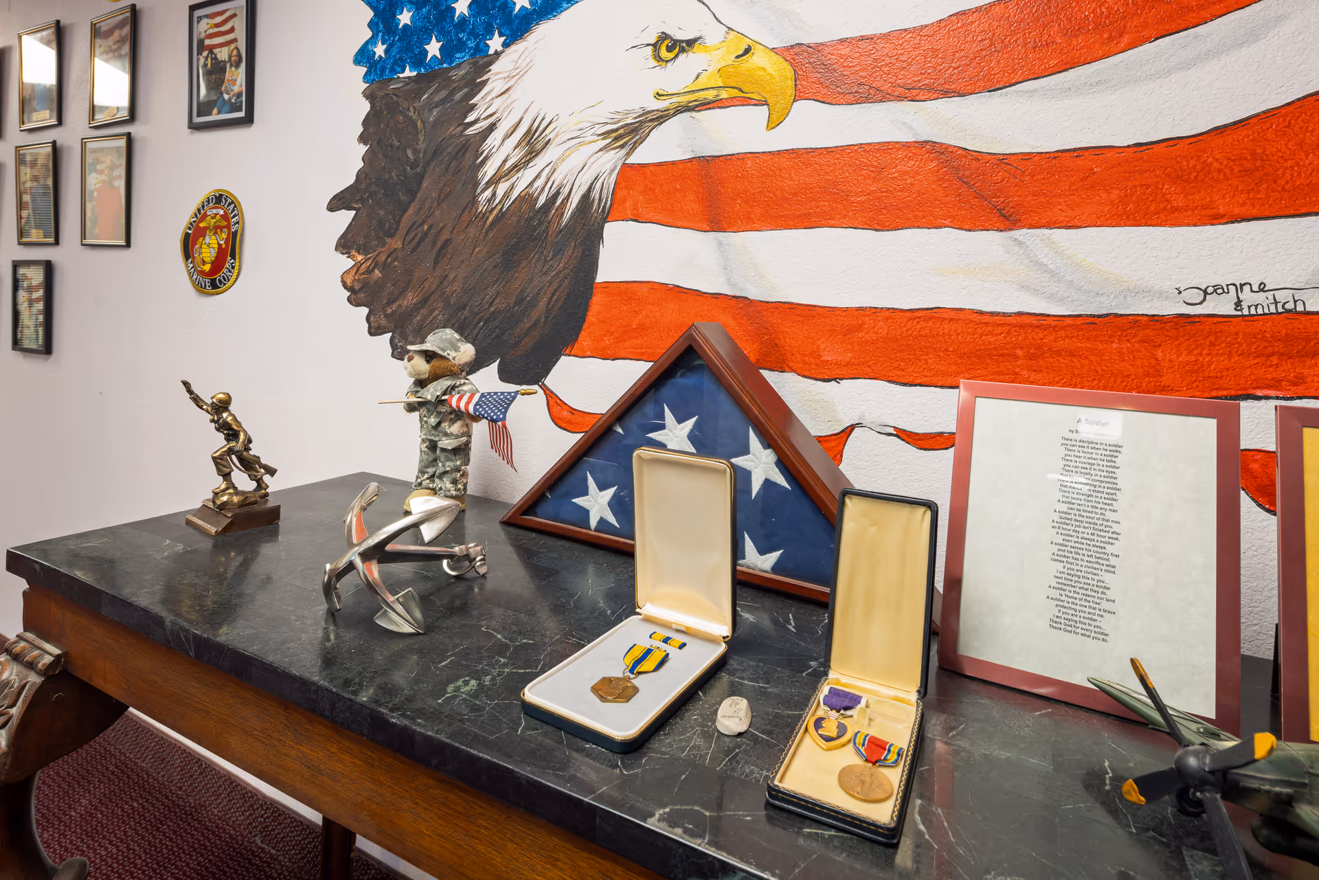 A display on a dark marble table featuring military memorabilia including two medal cases with medals, a folded American flag in a triangular wooden case, a small soldier figurine holding an American flag, a bronze statue of a soldier, an anchor sculpture, and a model airplane. Behind the table is a wall mural of a bald eagle with an American flag background and framed pictures and plaques on the adjacent wall.