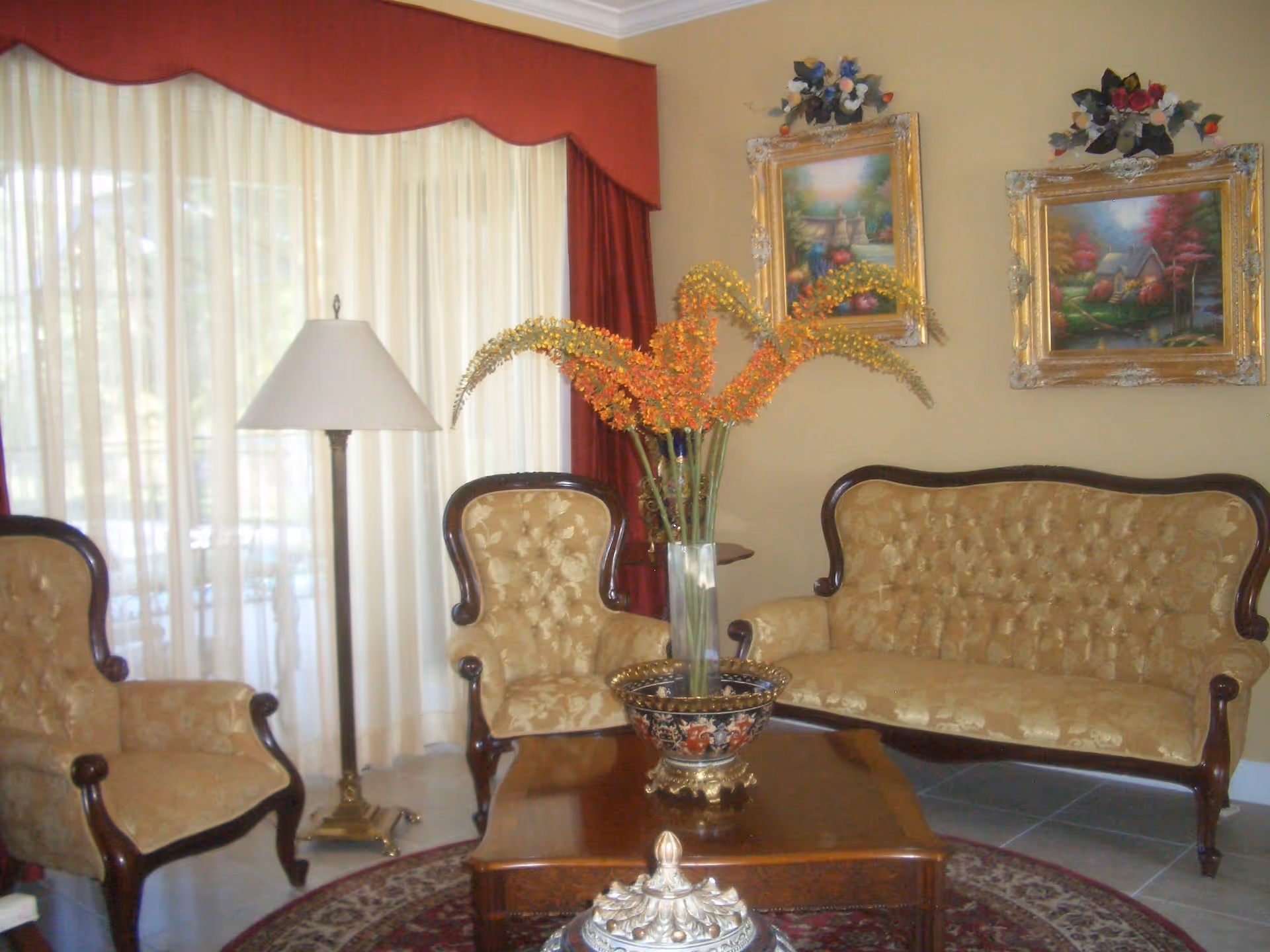 Decorative living room with tufted gold-upholstered sofa and chairs around a wooden coffee table topped with a tall vase of orange flowers, a floor lamp, framed paintings, and sheer curtained windows with a red valance.