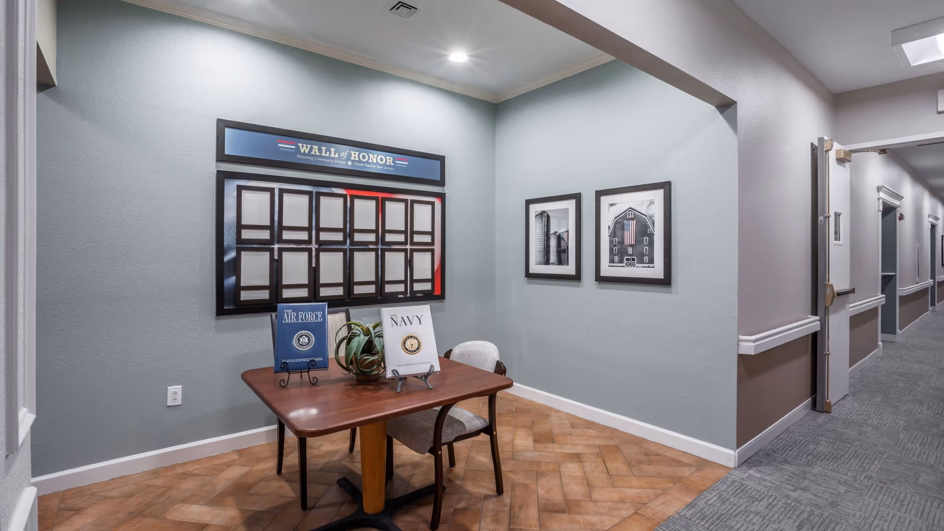 A hallway corner in a senior living facility with a small wooden table and two chairs. On the wall above the table is a 'Wall of Honor' display with empty frames. Two framed black and white photographs hang on the adjacent wall. The hallway extends to the right with doors and handrails along the walls.