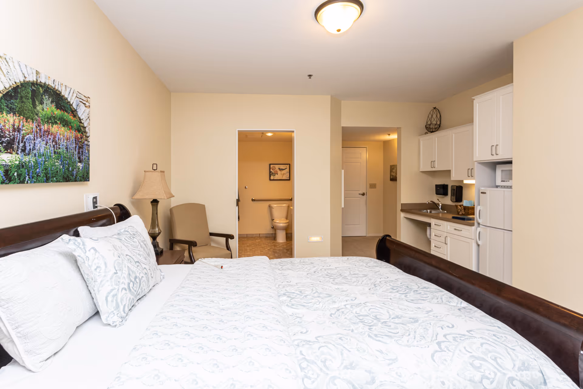 Well-lit senior living studio with a bed in the foreground, a chair and lamp, a kitchenette to the right, and a bathroom visible through an open doorway.
