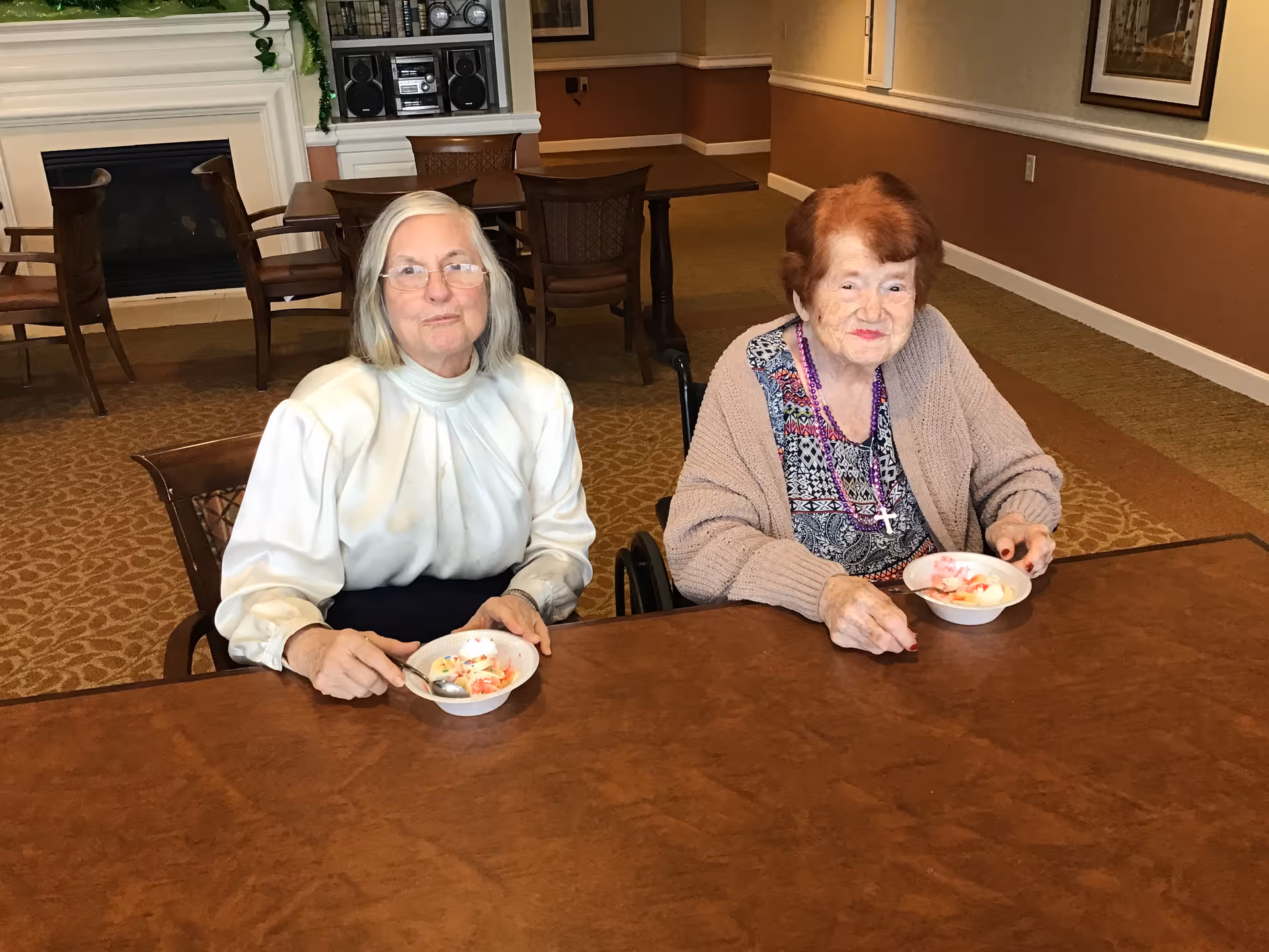 Two elderly women sit at a table eating from bowls in a communal dining area.