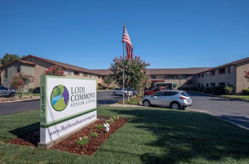 Exterior view of Lodi Commons Senior Living facility with a large sign displaying the facility name and website. The building is a two-story structure with beige walls and a brown roof. Several cars are parked in the driveway, and an American flag is flying on a flagpole near the entrance. The sky is clear and blue.