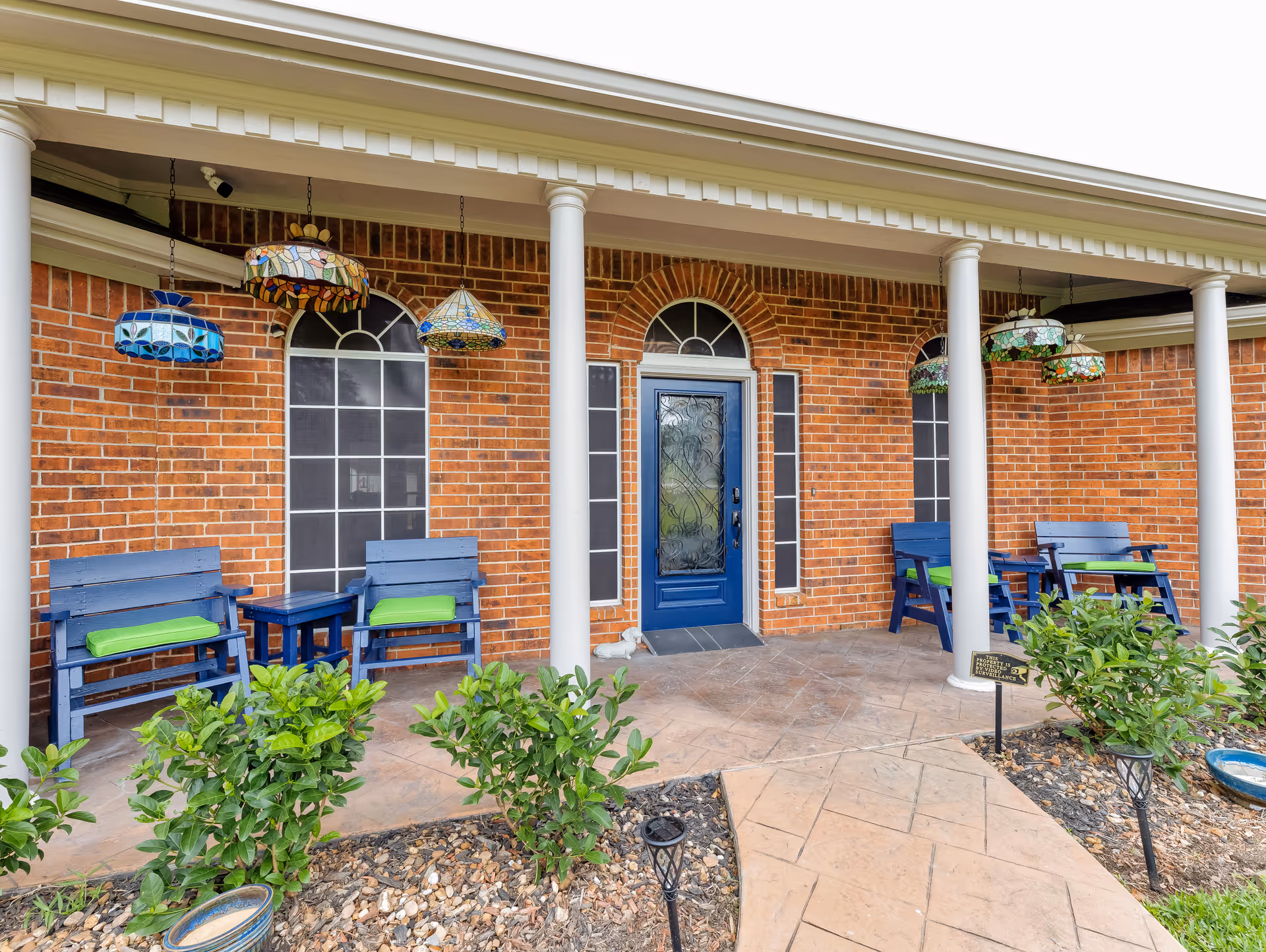 Brick-front covered porch with a blue decorative door, white columns, hanging stained-glass lamps, and blue bench seating with green cushions.