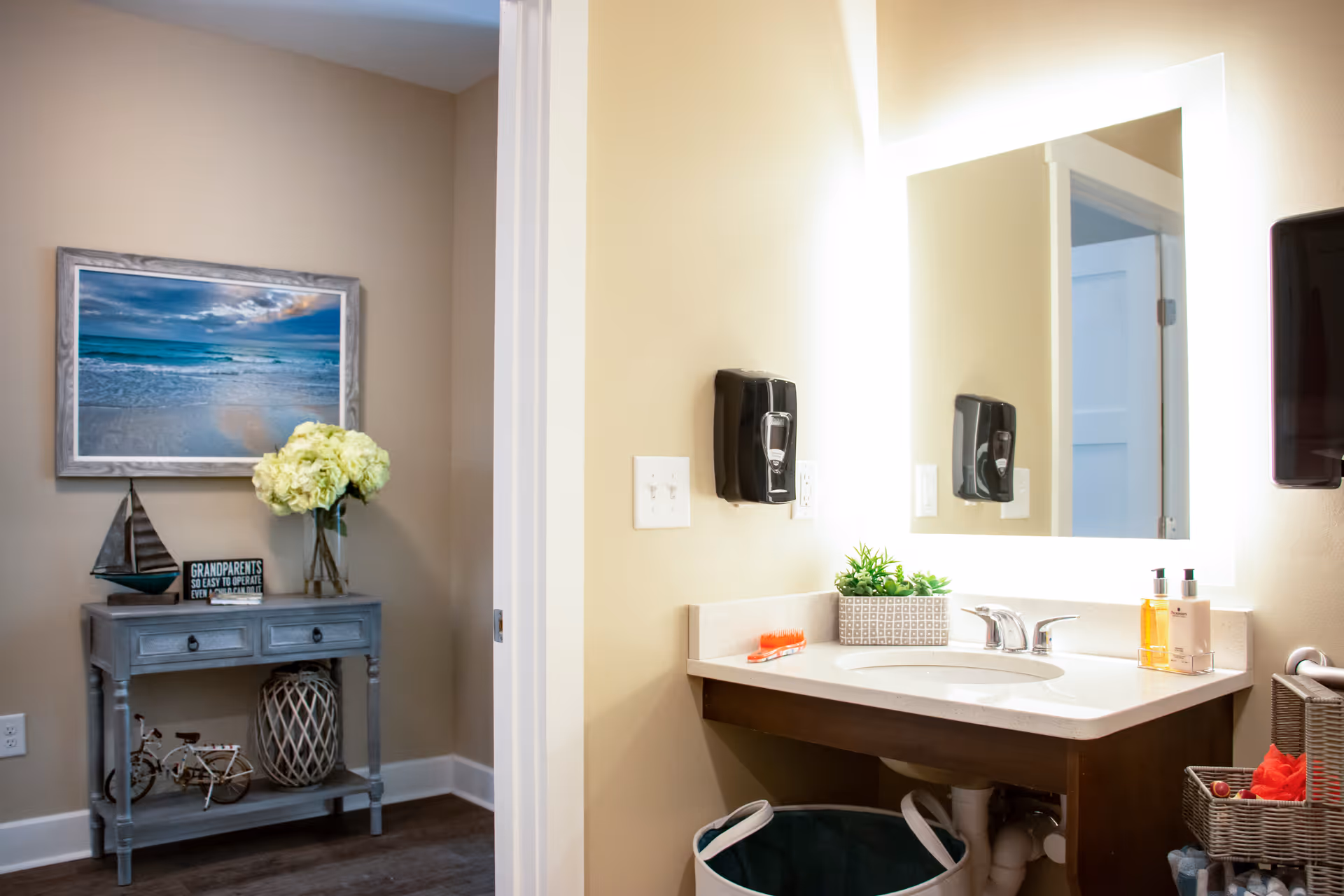 A bathroom vanity area with a lighted mirror, a sink with faucet, soap dispensers, a small potted plant, and a hairbrush on the counter. To the left, there is a small gray table with decorative items including a model sailboat, a vase with flowers, a small bicycle figurine, and a framed picture of a beach scene hanging on the wall above it.