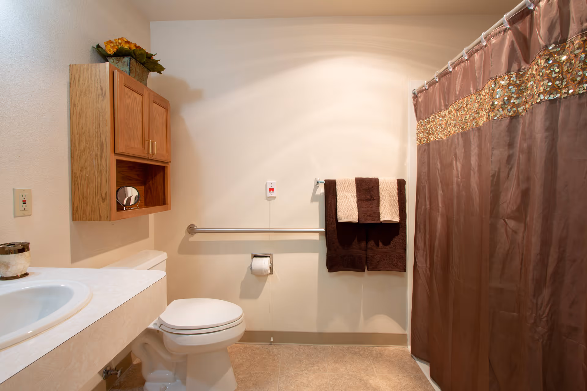 A clean bathroom with a white toilet, a sink with a countertop, a wooden cabinet mounted on the wall, a towel rack with brown and beige towels, and a shower with a brown curtain featuring a decorative gold and sequin band near the top.
