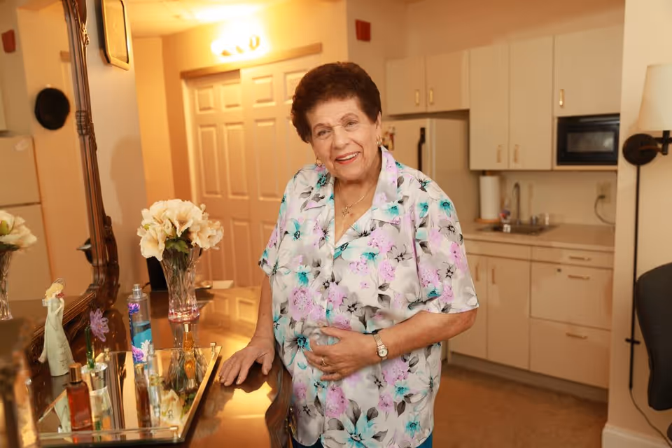 An elderly woman wearing a floral blouse stands smiling in a room with a dresser holding various perfume bottles and a vase of flowers. Behind her is a kitchenette with cabinets, a sink, and a microwave.