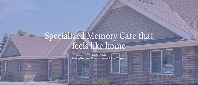 Exterior view of a brick building with multiple windows and a sloped roof under a clear blue sky. Text overlay reads 'Specialized Memory Care that feels like home. Family Owned. Serving the Dane County Community for 25 years.'