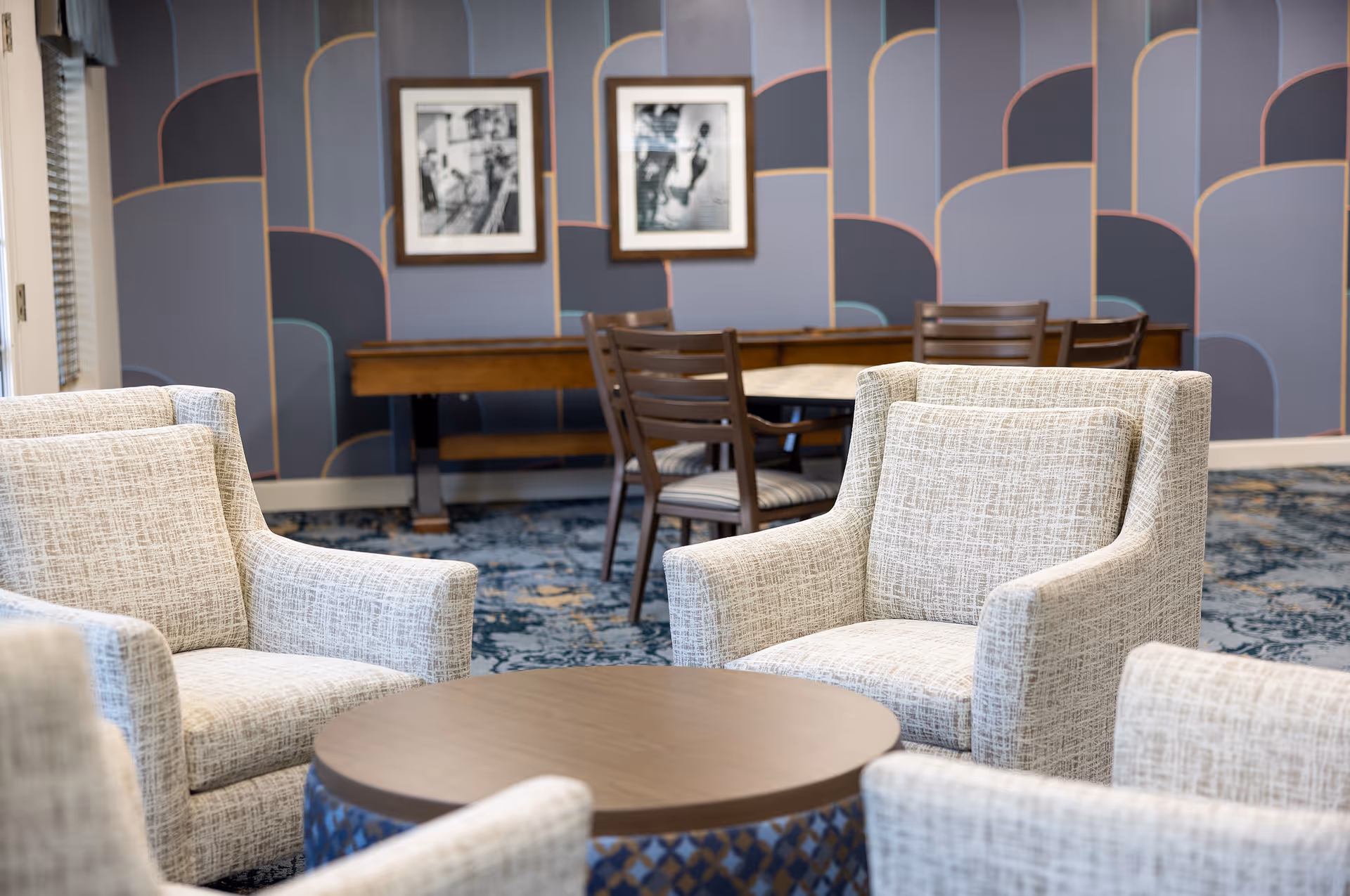 A cozy seating area with four beige patterned armchairs arranged around a round wooden table. In the background, there is a wooden table with four chairs and two framed black-and-white photos on a wall with a geometric blue and purple pattern.