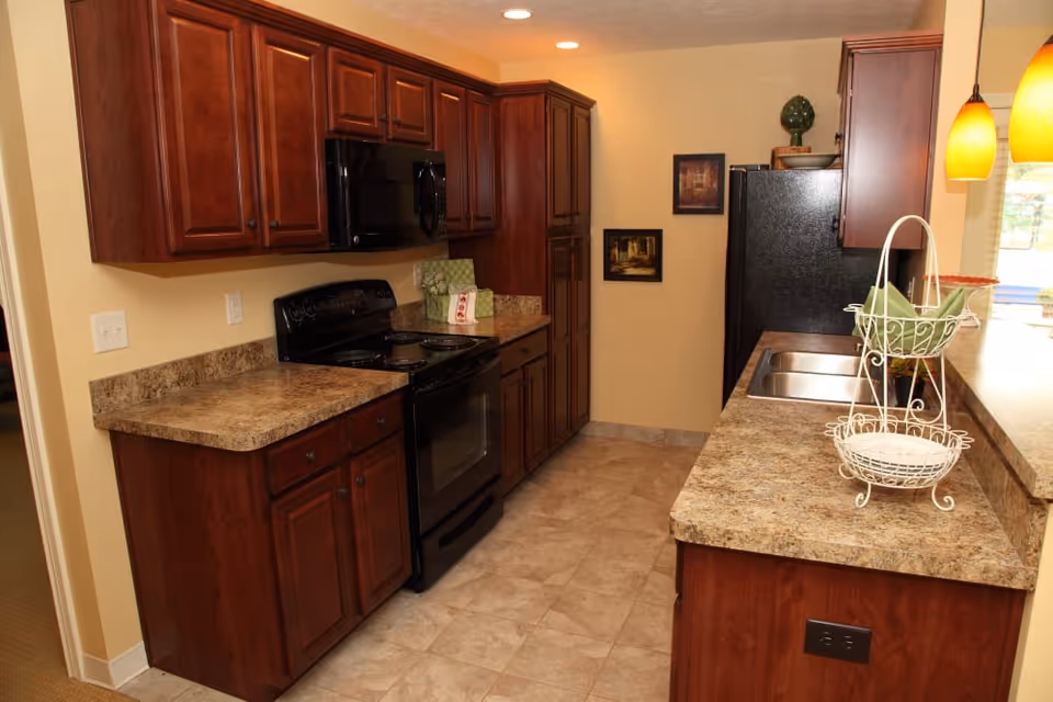 A kitchen with dark wood cabinets, a black stove and microwave, a double sink, and granite countertops. The floor is tiled, and there are two yellow pendant lights hanging above the counter. Two framed pictures hang on the wall near a black refrigerator.