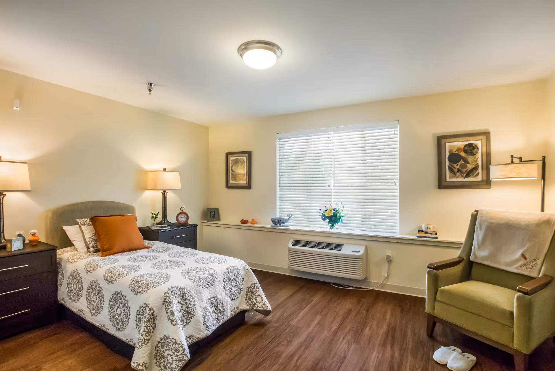 A cozy bedroom with a single bed covered in a white and gray patterned bedspread and an orange pillow. There are two dark wooden nightstands on either side of the bed, each with a lamp and small decorative items. A large window with white blinds lets in natural light. To the right, there is a green armchair with a Vineyard Place blanket draped over the back and a pair of white slippers on the floor. The room has wooden flooring and light-colored walls with framed artwork.