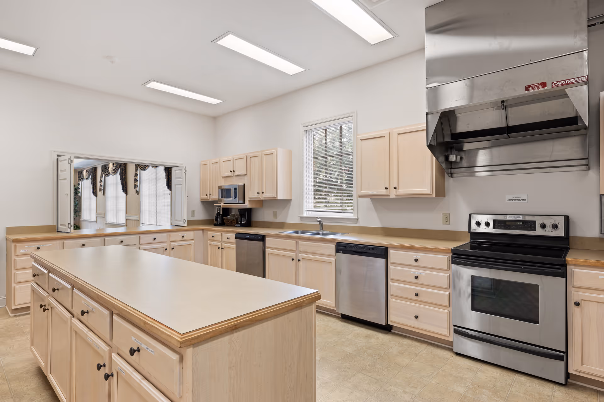 A spacious kitchen with light wood cabinets, a large central island, stainless steel appliances including a stove, dishwasher, and microwave, and a window above the sink letting in natural light. The kitchen has a pass-through window with a view into an adjoining room with large windows and draped curtains.