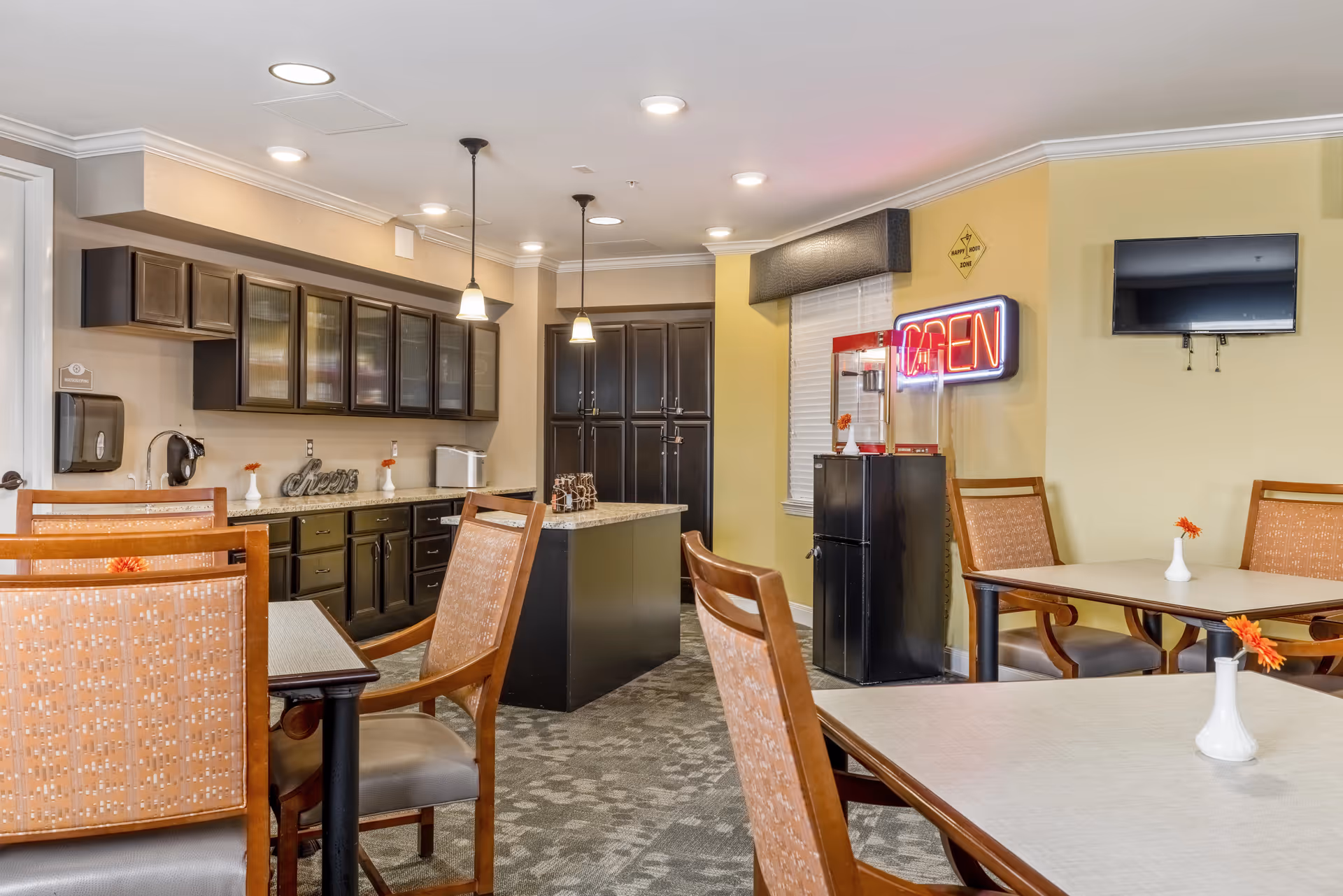 Communal dining area with tables and chairs, a kitchenette with dark cabinets and an island, and a neon "OPEN" sign on the wall.