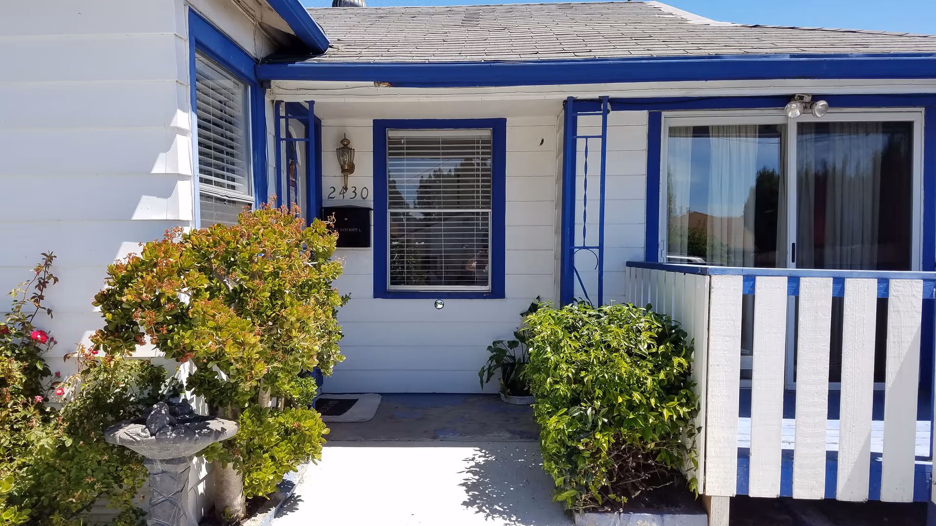 Front porch of a white house with blue trim, featuring a window with blinds, a door with the number 2430, a small garden with green bushes and flowers, and a white and blue railing on the right side.