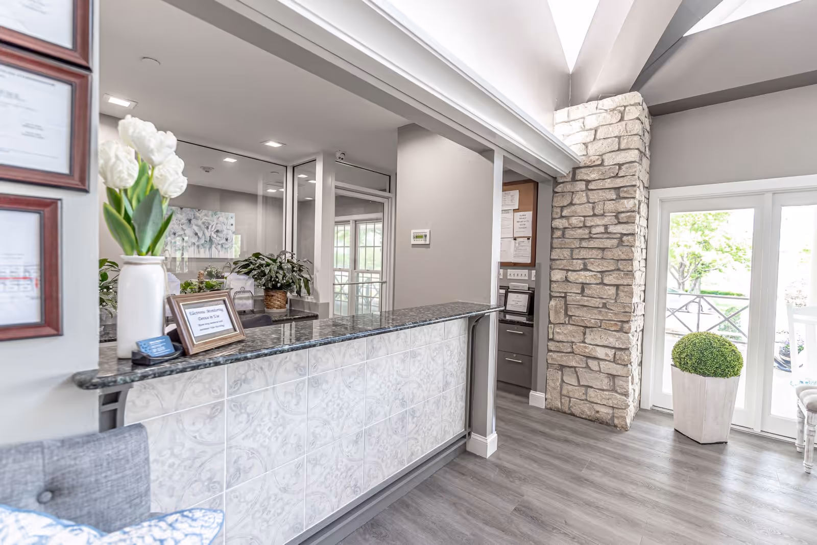 Reception area with a granite countertop and decorative tiled front, a vase with white flowers, framed certificates on the wall, potted plants, a stone accent wall, and glass doors leading outside with a potted shrub visible.