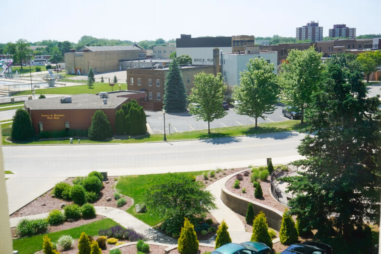 View of a small town area with a landscaped garden in the foreground, a road, and several buildings including a Farmers & Merchants State Bank building and other commercial buildings in the background under a clear sky.
