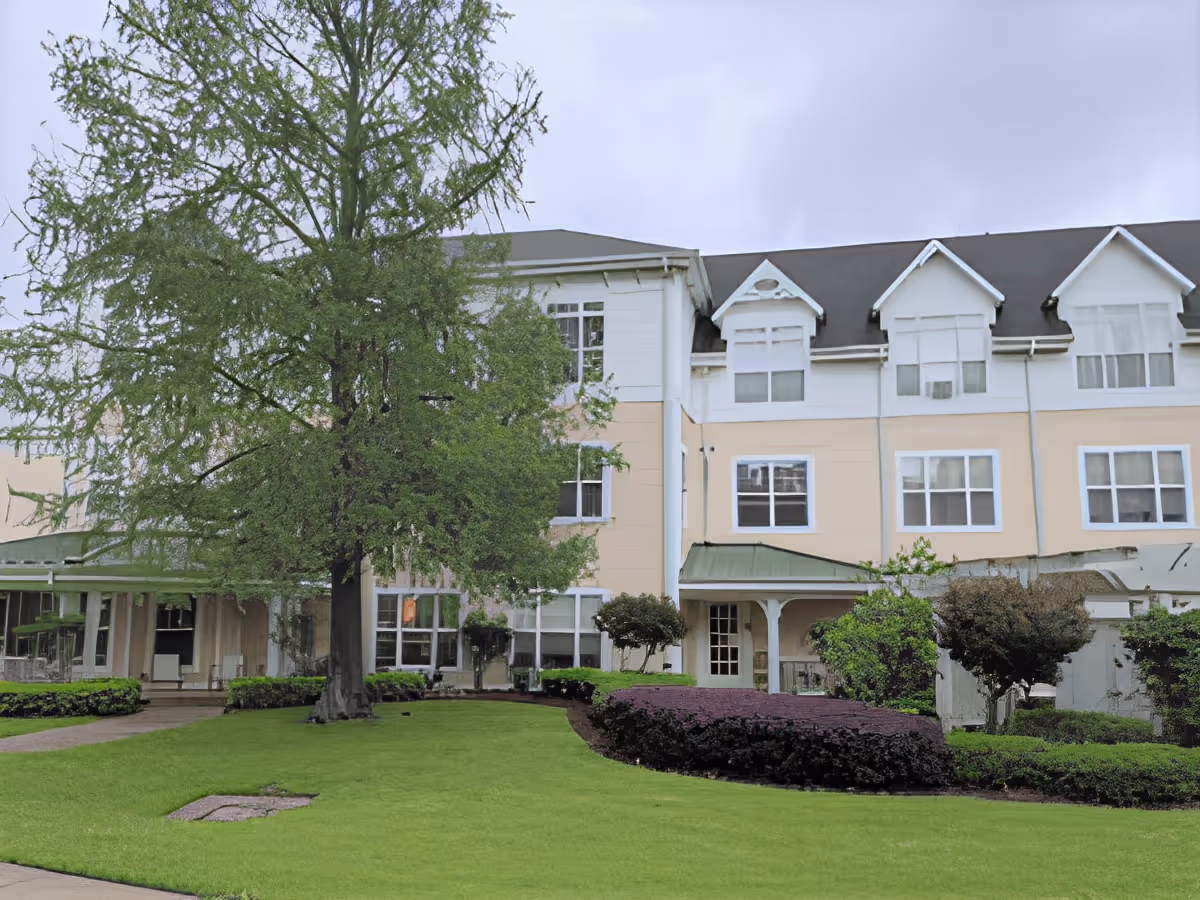 Exterior view of a multi-story senior living facility building with beige and white walls, multiple windows, a green lawn, bushes, and a large tree in front under a cloudy sky.