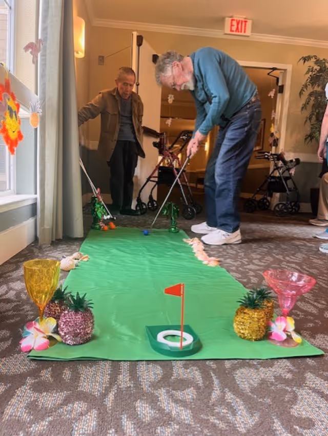 Two elderly men playing indoor mini golf on a green putting mat decorated with small pineapples, tropical flowers, and colorful cups in a well-lit room with carpeted floor and an exit sign above a door in the background.