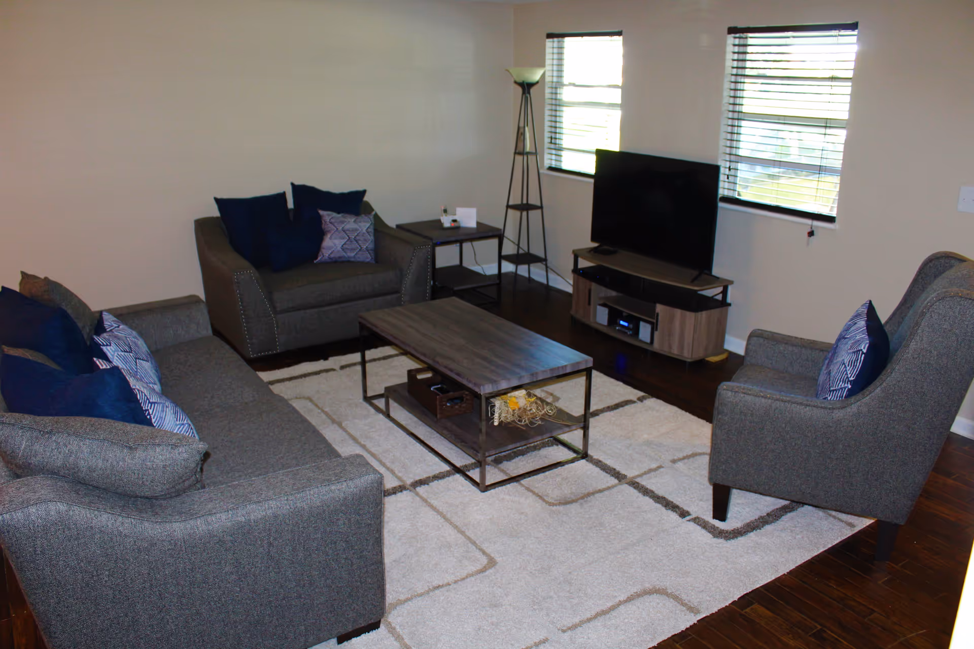 A living room with a gray sofa, a matching armchair, and a loveseat, all with blue and patterned throw pillows. There is a rectangular wooden coffee table on a light-colored area rug with a geometric pattern. A TV is placed on a wooden TV stand against the wall, with two windows behind it. A tall floor lamp stands next to the TV stand.