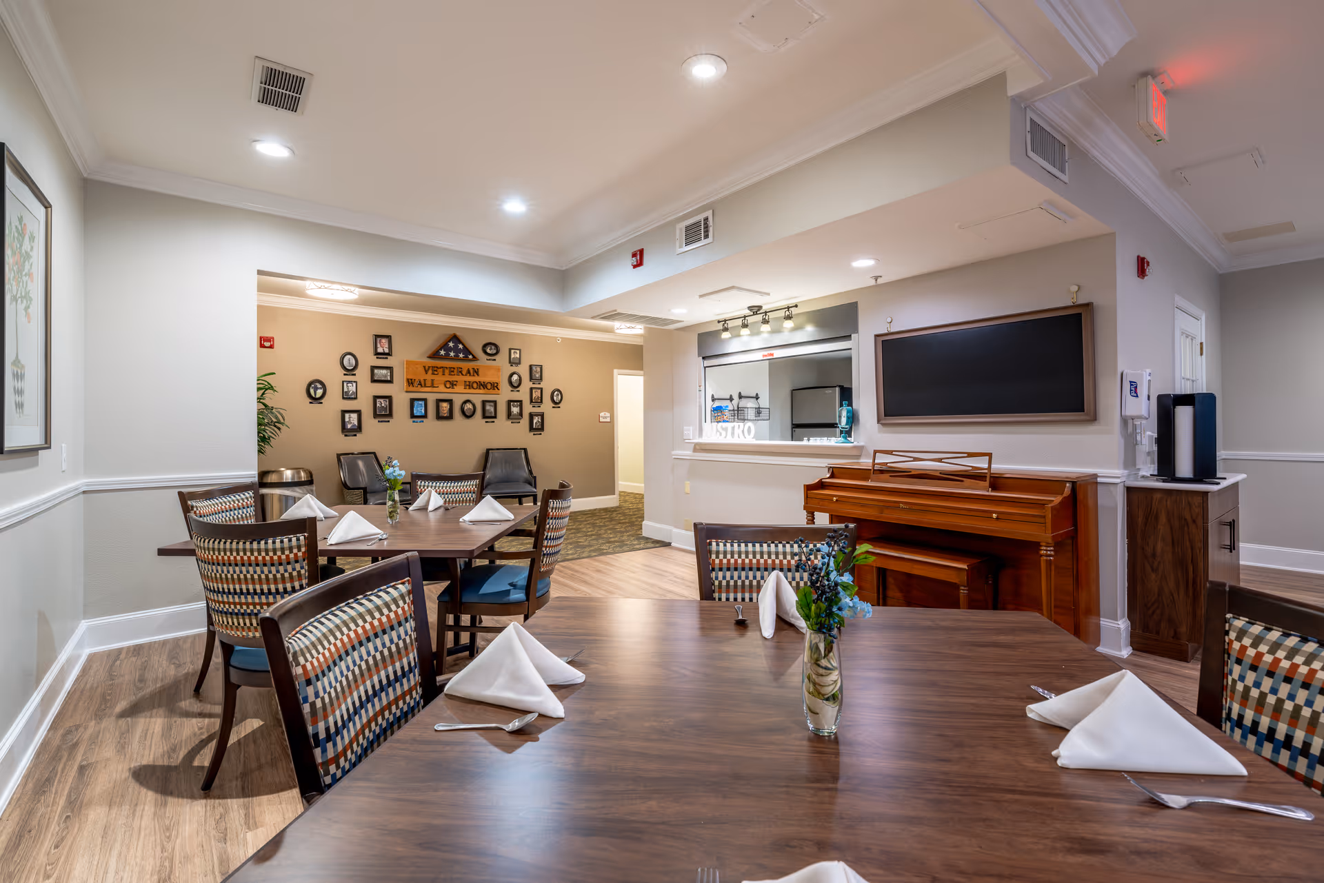 A dining area in a senior living facility with wooden tables set with white folded napkins and silverware. Chairs with patterned upholstery surround the tables. A wooden piano is against the wall, and a blackboard hangs above it. In the background, there is a 'Veteran Wall of Honor' with framed photos and memorabilia. The room has soft lighting and wood flooring.