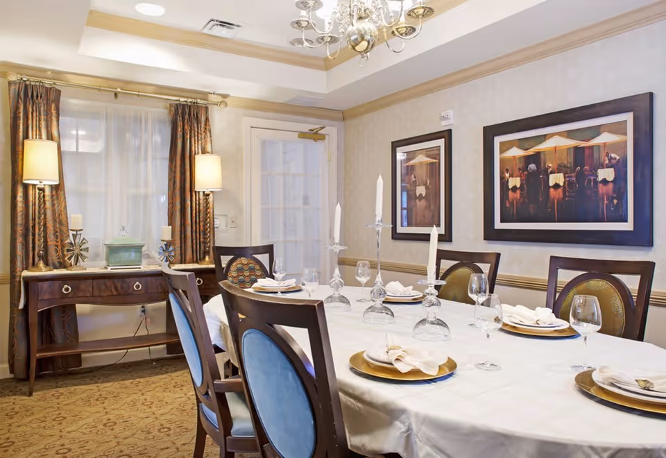 A formal dining room with a long table covered with a white tablecloth, set with plates, napkins, wine glasses, and tall candlesticks. The room has elegant wooden chairs with blue upholstery, a side table with two lamps and decorative items, patterned curtains, and framed artwork on the wall.