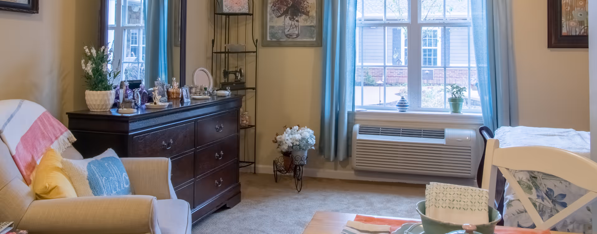 A cozy living room area with a beige armchair adorned with colorful pillows and a throw blanket. A dark wooden dresser with various decorative items and a potted plant sits against the wall. A window with blue curtains lets in natural light, with a small plant and decorative vase on the windowsill. A small table with a floral arrangement and a white chair with a floral cushion are also visible.