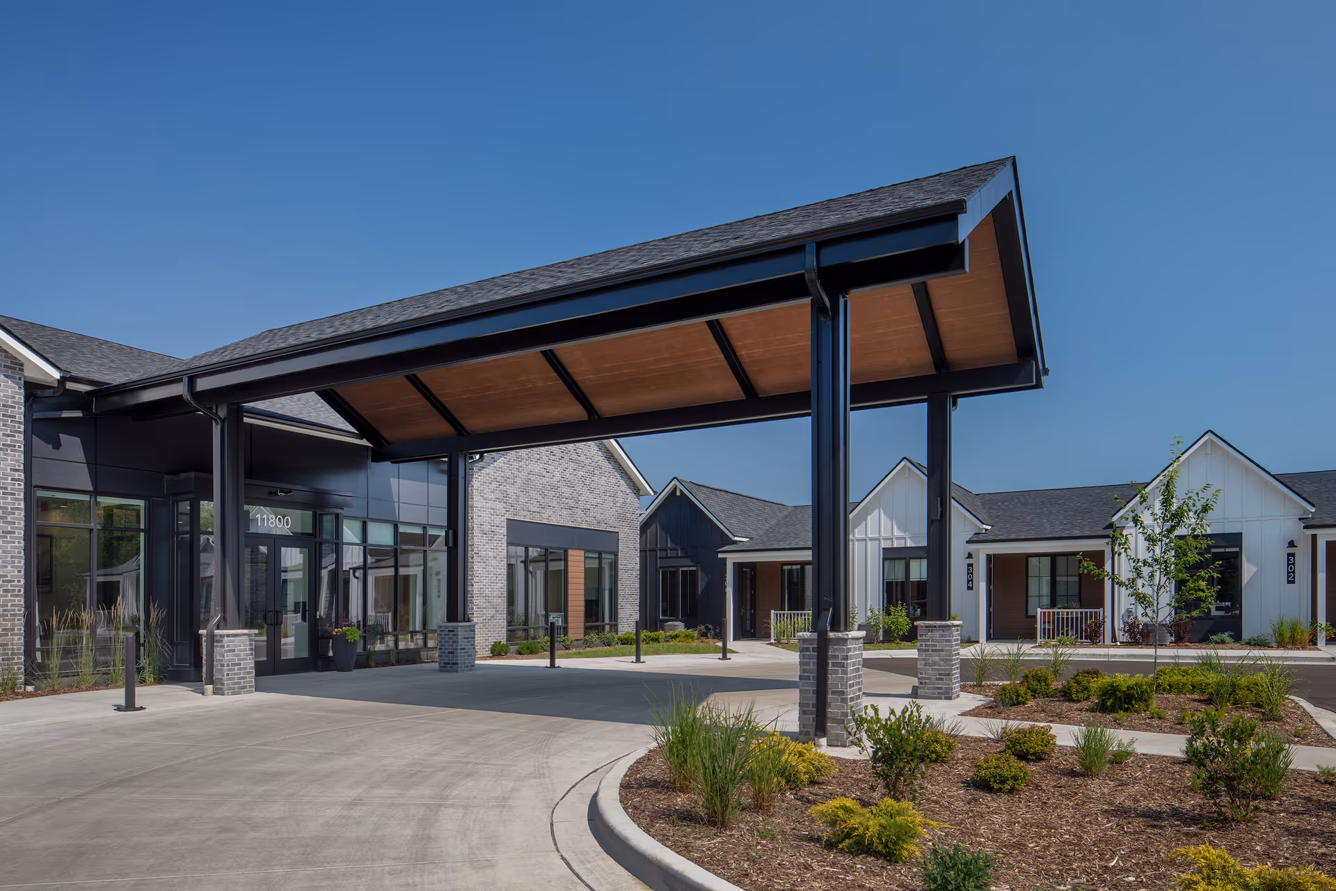 Exterior view of The Woods - A GreatLife Community senior living facility showing a covered entrance with a modern roof supported by black pillars, surrounded by landscaped greenery and multiple residential-style buildings under a clear blue sky.