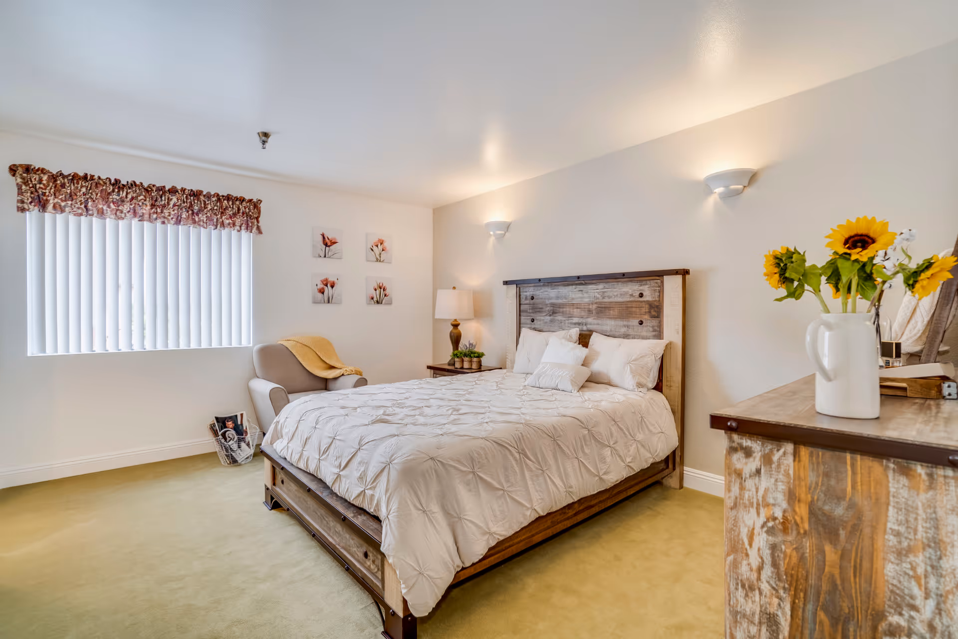 A cozy bedroom with a wooden bed frame and a white comforter. There is a beige armchair with a yellow blanket near a window with vertical blinds and a floral valance. On the wall above the chair are four framed floral pictures. A wooden nightstand with a lamp and small plants is next to the bed. A wooden dresser with a white vase holding sunflowers is visible in the foreground.
