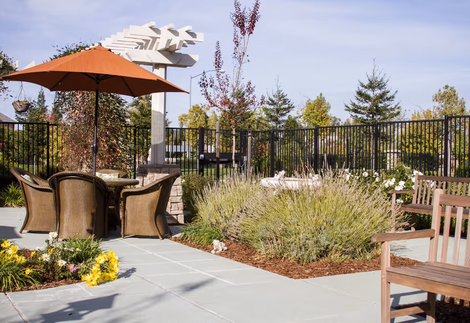 Outdoor patio area with a round table and four wicker chairs under a large orange umbrella. There are wooden benches, various plants, flowers, and shrubs surrounding the paved patio. A black metal fence encloses the area with trees and greenery visible beyond it under a clear sky.