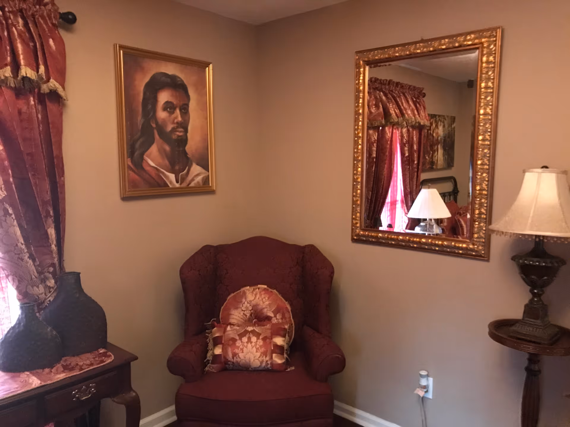 A cozy corner of a room featuring a burgundy upholstered armchair with decorative pillows, a wooden side table with two dark vases, a large ornate gold-framed mirror reflecting a lamp and window with burgundy curtains, a framed portrait of a man with long hair and beard on the wall, and a table lamp on a small round wooden table.