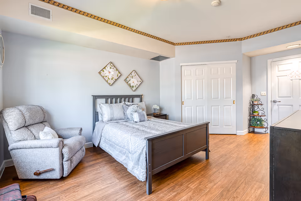 A bright and tidy bedroom with a wooden bed frame, neatly made bed with gray and white bedding, a gray recliner chair, two floral wall decorations above the bed, a wooden nightstand with a small flower arrangement, a closet with white sliding doors, and a small black metal shelf with plants and decor near a white door.