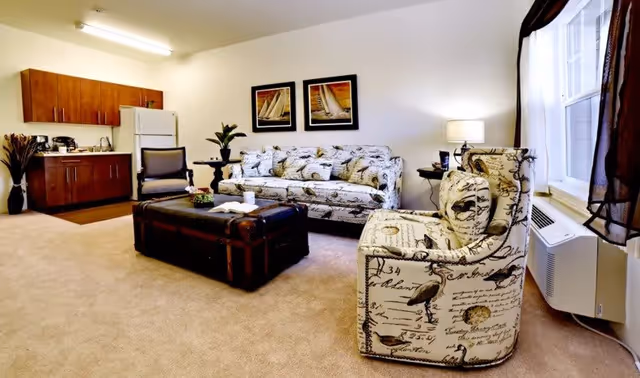 A cozy living room area in a senior living facility featuring a patterned armchair and matching sofa with bird and script designs, a dark wooden coffee table, a side table with a lamp, and a kitchenette with wooden cabinets, a refrigerator, and a sink. The room has beige carpeting and a window with dark curtains.