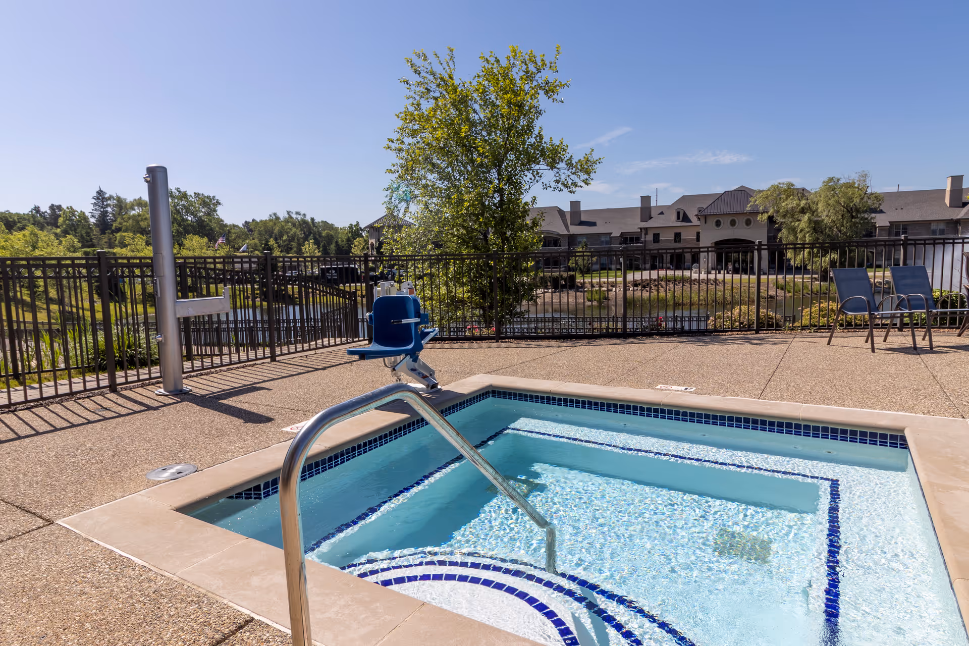 Outdoor spa pool with a handrail and blue pool lift on a patio, overlooking fenced grounds and a residential building.