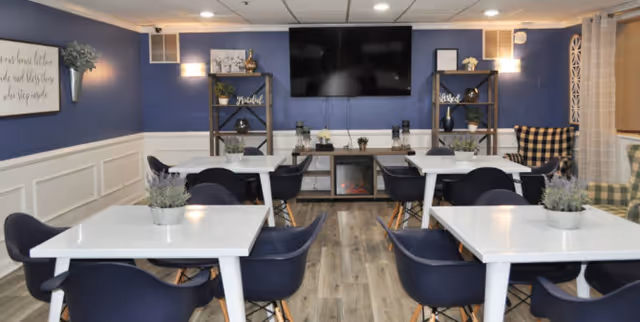A cozy common area with four white tables each surrounded by four dark blue chairs. Each table has a small potted plant centerpiece. The room has blue walls with white wainscoting, wood flooring, and a large flat-screen TV mounted on the wall. There are two shelving units with decorative items and a fireplace below the TV. In the corner, there are two plaid armchairs, one in black and white and the other in green and white. Soft lighting fixtures are mounted on the walls.