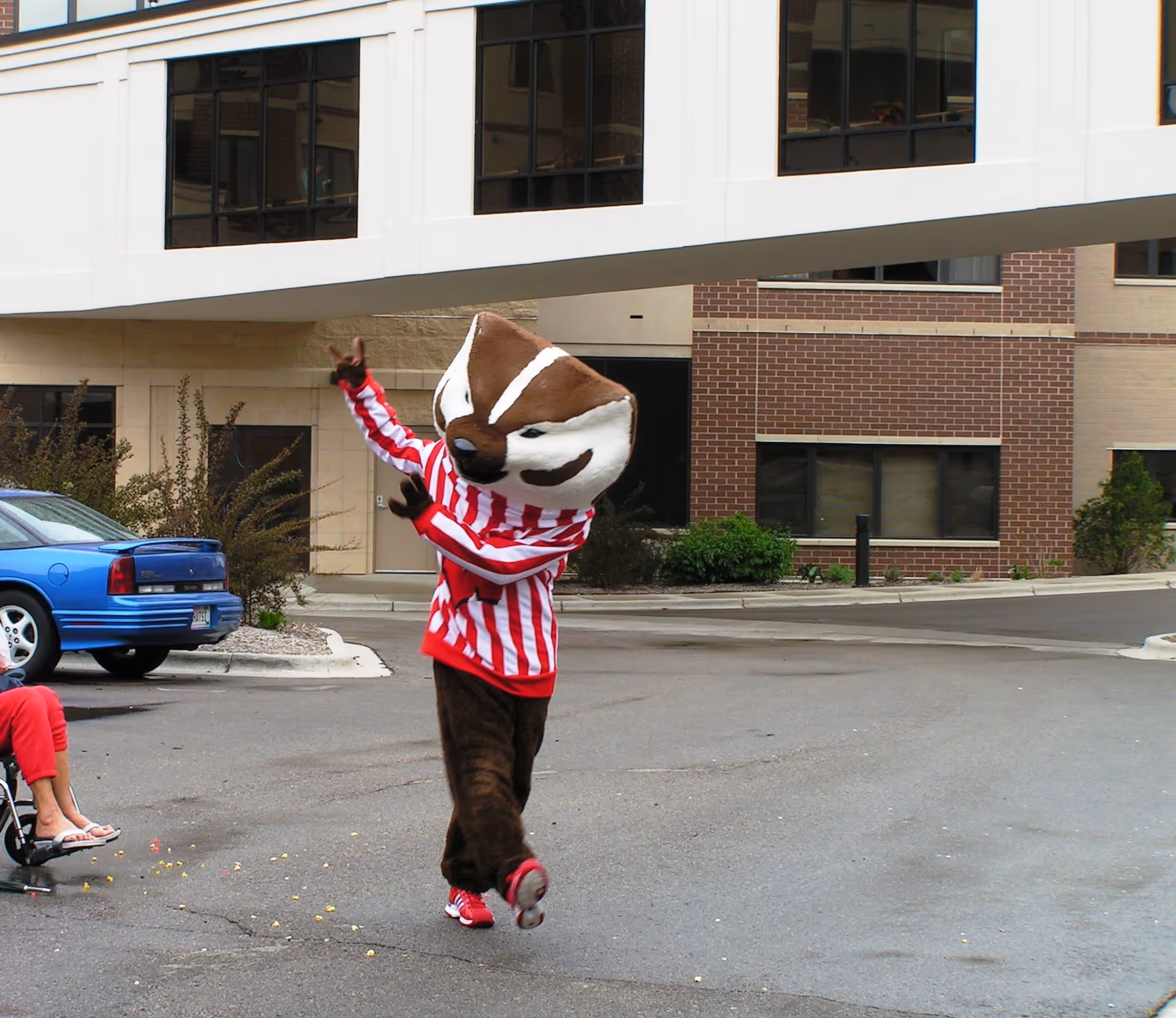 A person in a large brown-and-white mascot costume wearing a red-and-white striped shirt dances in a parking lot in front of a brick building.