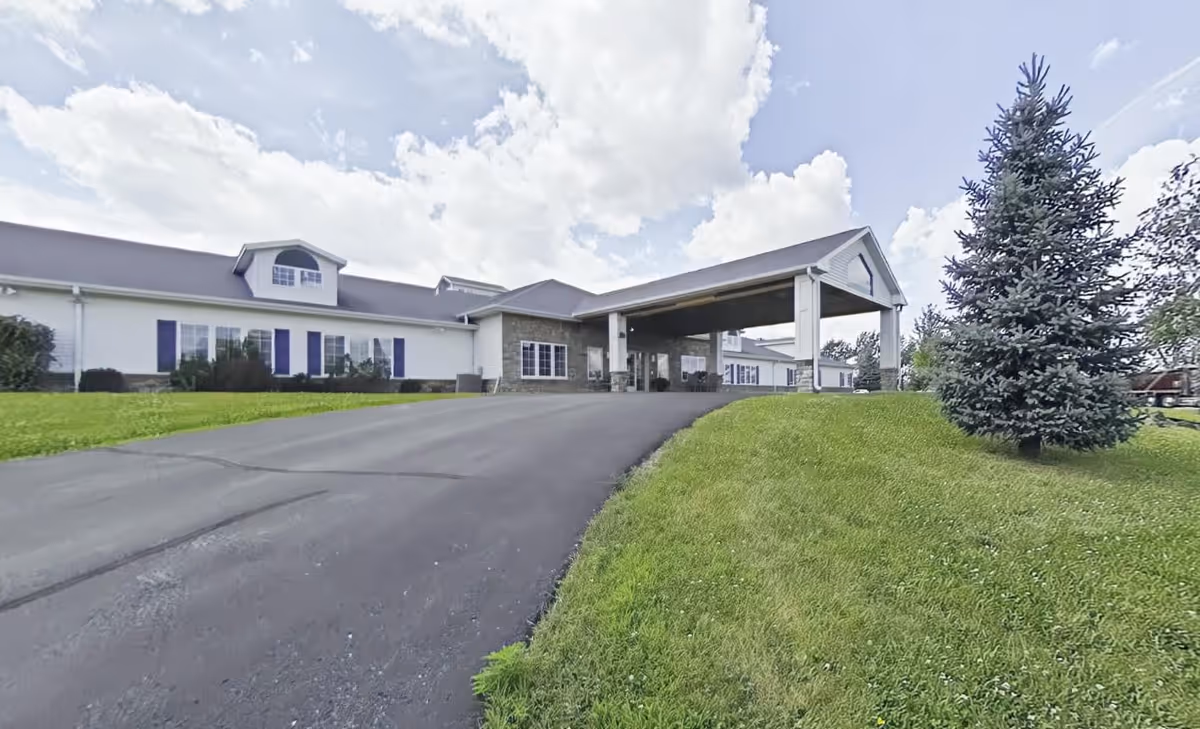 Exterior view of Charleston House Memory Care facility showing a paved driveway leading up to a covered entrance. The building is white with dark shutters and has a well-maintained lawn with a large evergreen tree on the right side. The sky is partly cloudy.