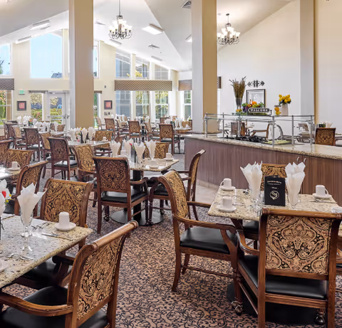 A spacious and well-lit dining room with multiple tables and chairs arranged neatly. Each table is set with white napkins folded elegantly, cups, and glasses. The room features large windows allowing natural light to fill the space, chandeliers hanging from the ceiling, and a buffet area with decorative plants and a welcome sign.