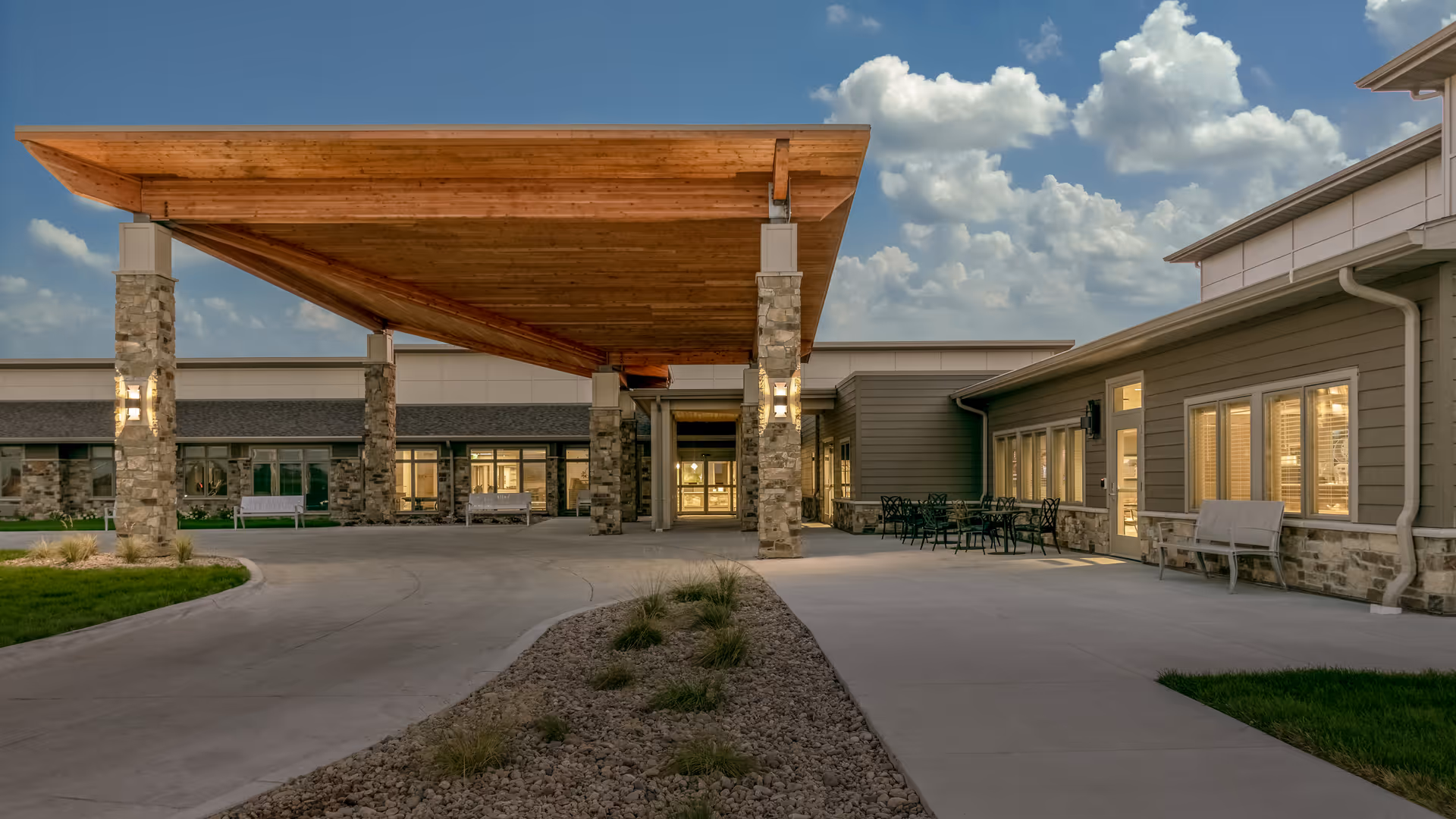 Entrance of Brookestone Gardens with a large wooden canopy supported by stone columns over a driveway leading to the well-lit building.
