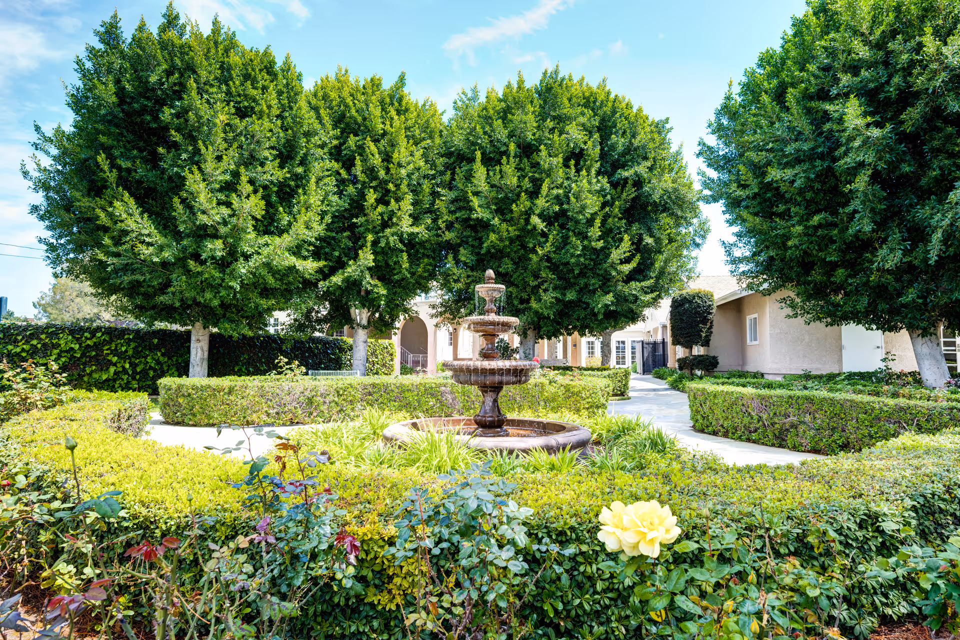 A landscaped outdoor garden area with a tiered water fountain in the center, surrounded by neatly trimmed hedges, flowering plants, and several large green trees. A paved walkway curves around the garden leading to beige buildings in the background under a clear blue sky.