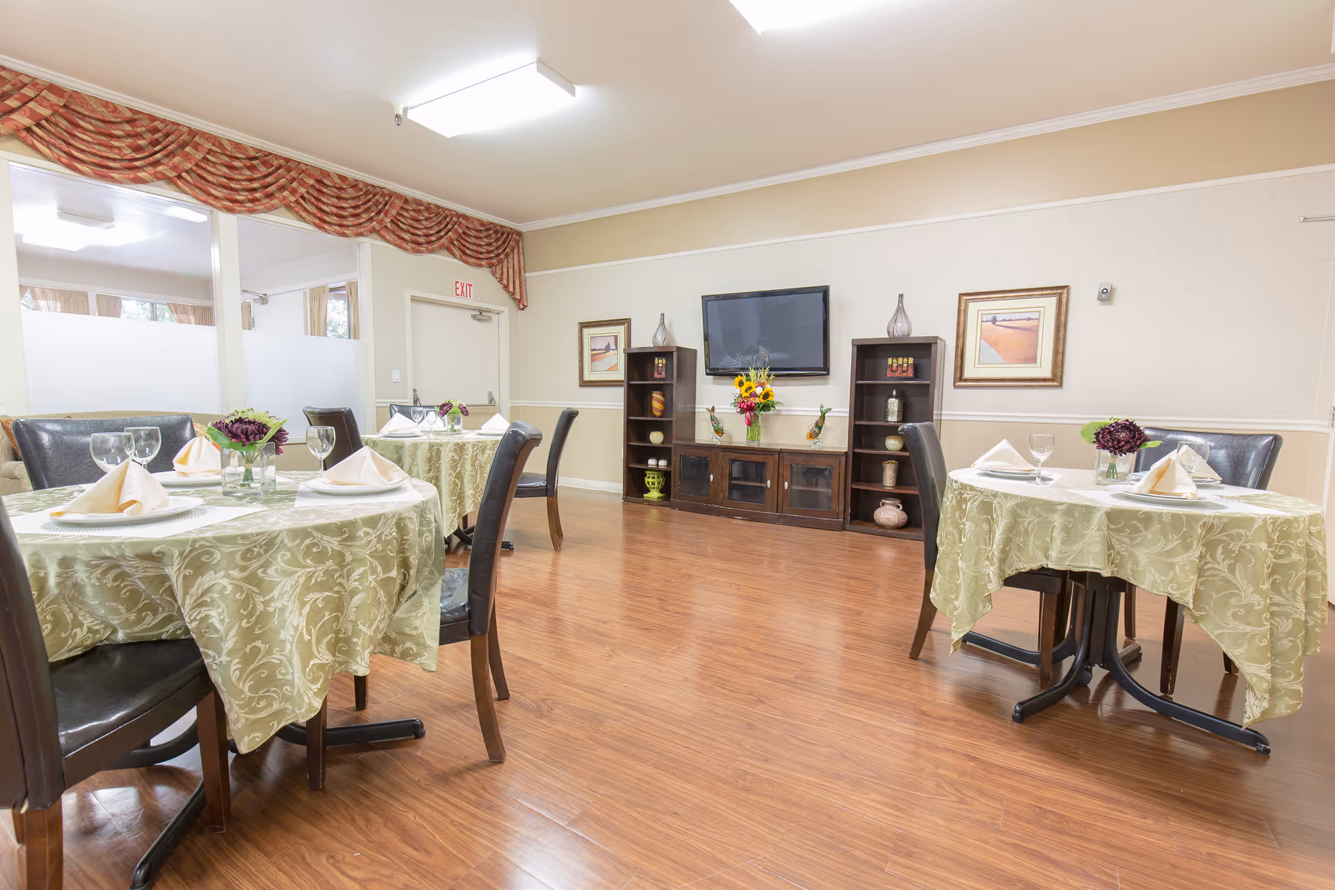 A dining room with round tables covered in green patterned tablecloths, each set with folded napkins, glassware, and small floral centerpieces. The room has wooden flooring, a wall-mounted TV, decorative shelves with vases, framed artwork, and an exit door with a red exit sign above it.