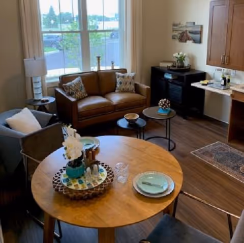 Cozy assisted-living interior with a round dining table in the foreground, a sofa and armchair by a window, and a small kitchenette and cabinetry.