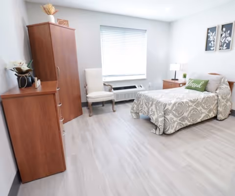 A bright and clean bedroom with light gray walls and flooring. The room features a single bed with a patterned bedspread and a green pillow that says 'blessed'. There is a wooden nightstand with a lamp and a small plant, a wooden dresser with decorative items, a wooden wardrobe, and a white upholstered chair near a window with blinds.