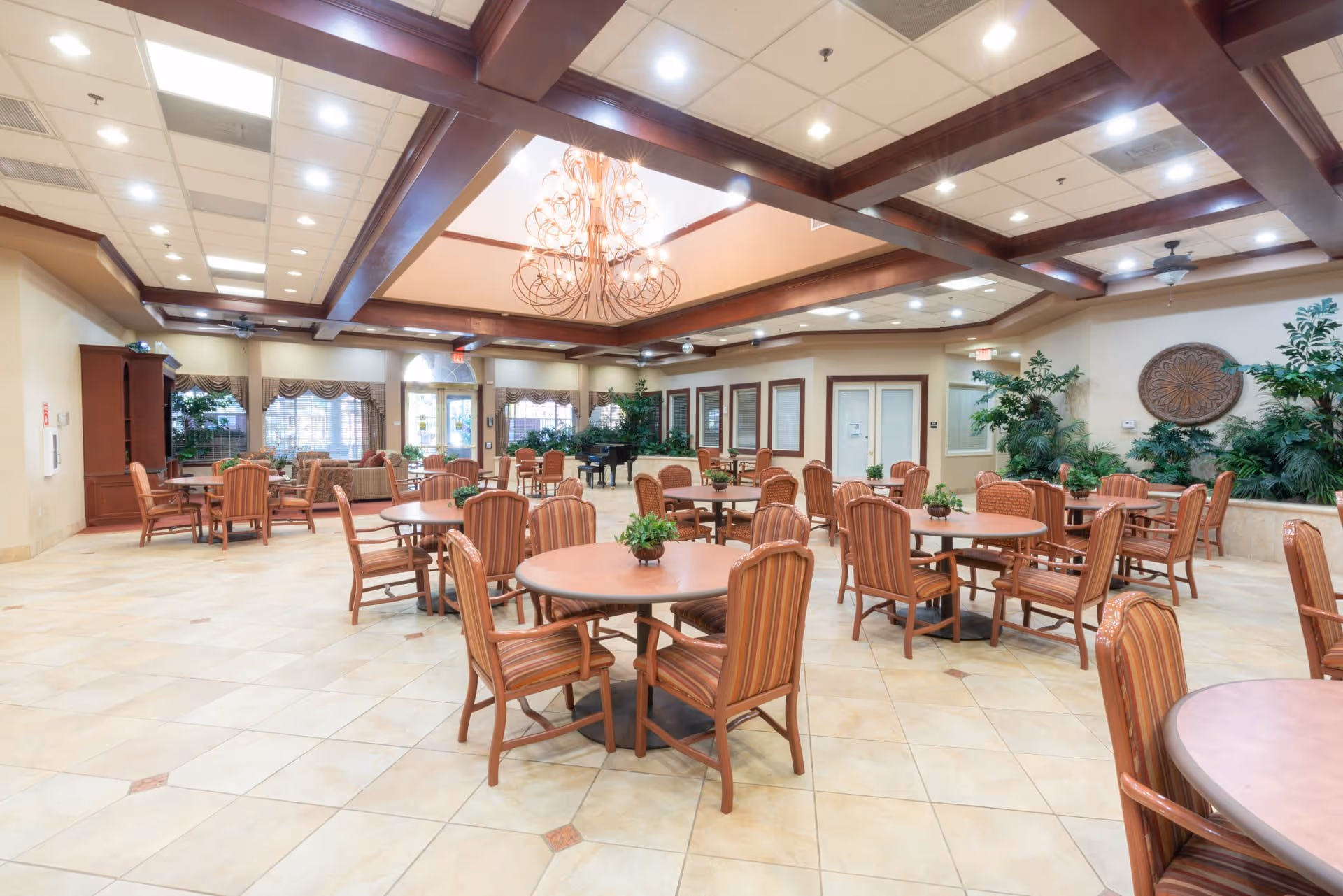 Spacious and well-lit common area with multiple round tables and striped cushioned chairs arranged neatly. The room features a high ceiling with wooden beams and a decorative chandelier. There are large windows with valances, several potted plants, and a piano in the background, creating a welcoming and comfortable atmosphere.