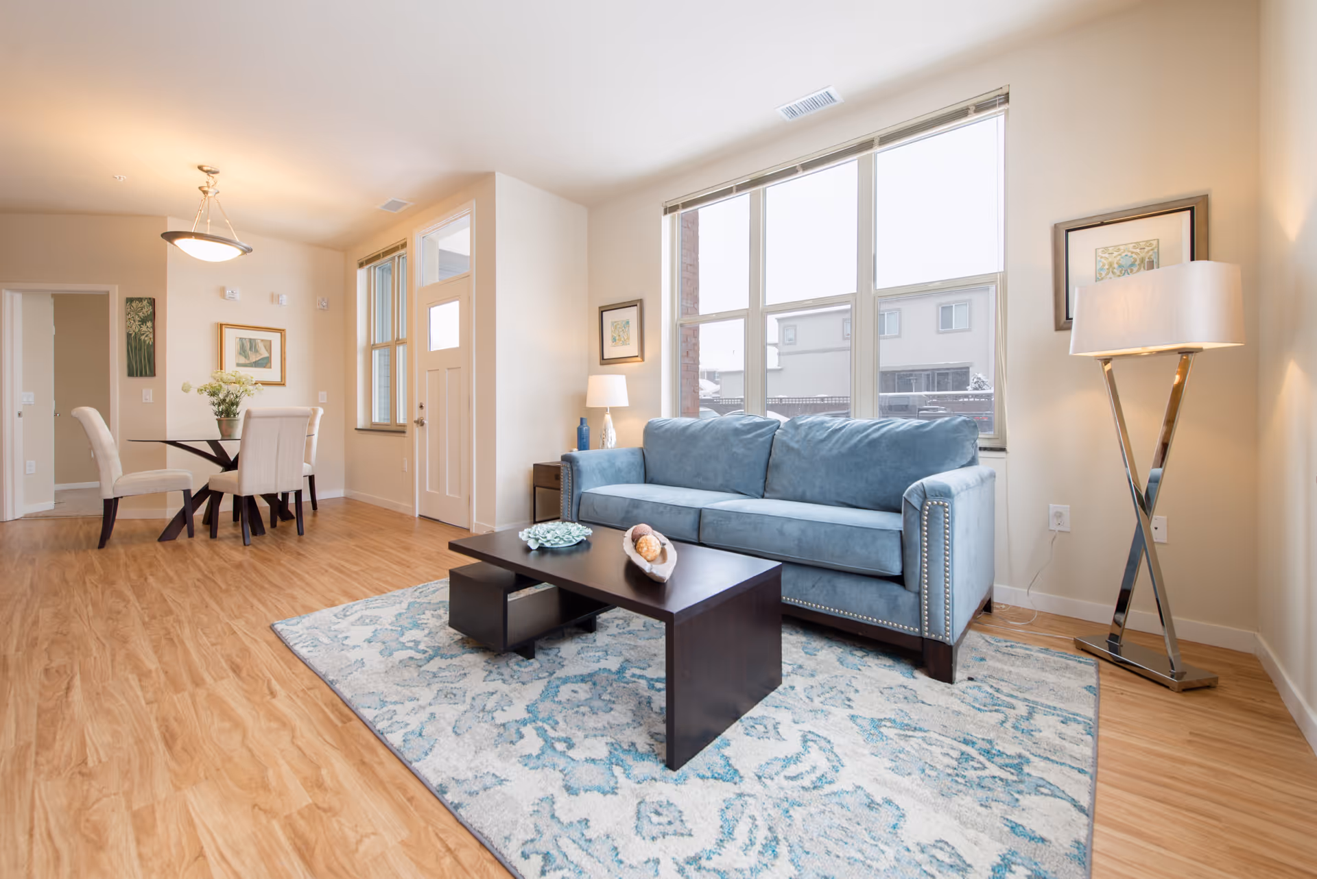 Bright living room with a large window, a blue upholstered sofa, a dark wooden coffee table on a patterned rug, a floor lamp with a white shade, and a dining area with a round glass table and four beige upholstered chairs. Light wood flooring and neutral-colored walls with framed artwork complete the space.