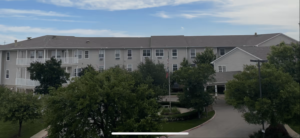 Exterior view of a large, multi-story senior living facility building with beige siding and multiple windows. The building is surrounded by green trees and a driveway curves in front of the entrance. The sky is partly cloudy.