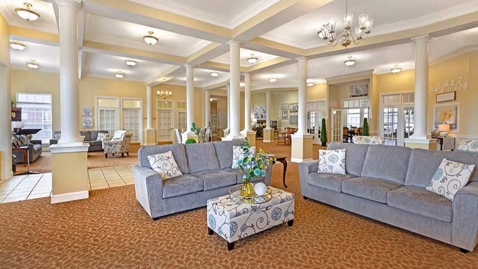 Bright, open senior living common room with gray sofas, a patterned ottoman, columns, and chandeliers.